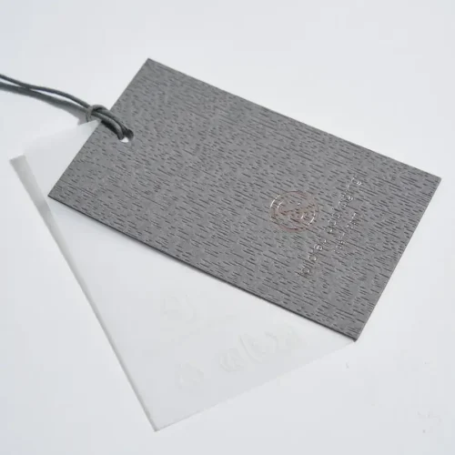 Coppia di cartellini Coein sovrapposti: una base in vero piallaccio di legno grigio con venature a rilievo e logo in lamina rame, abbinata a un sottostrato in carta pergamena semitrasparente